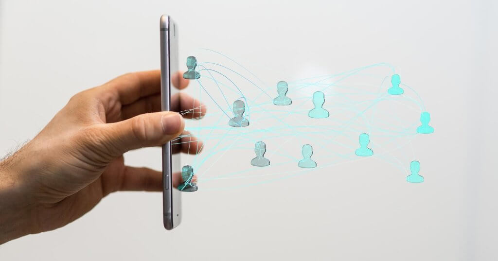 Mão segurando um smartphone na vertical com ícones de usuários conectados em rede, representando comunicação, compartilhamento de dados e conectividade digital.